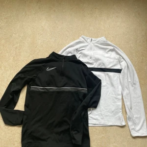 Nike half zip tröjor svart & vit - Två snygga Nike tröjor med half zip, en svart och en vit. Båda har lång ärm, broderad Nike-logga på bröstet och en kontrastfärgad rand över bröstet. Perfekta för träning eller chill. Materialet känns mjukt och sportigt.