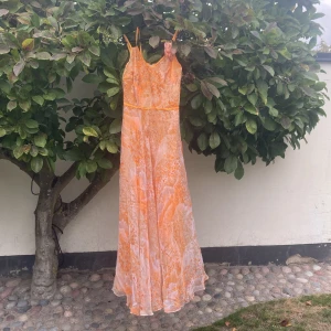 Lång blommig klänning i orange och rosa - Lång klänning med smala axelband i en mix av orange och rosa toner. Klänningen har blommigt mönster, v-ringning och en dekorativ blomma på axeln. Tillverkad i ett lätt, glansigt material med dragkedja i ryggen och markerad midja för en flowy siluett.