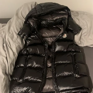 Svart dunväst  - Snygg svart dunväst med glansig finish och klassisk logga på bröstet. Västen har huva, två dragkedjefickor och quiltad design. Perfekt för lager-på-lager och håller dig varm under kyliga dagar.