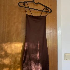 Brun satin klänning från H&M Divided - Snygg brun kortklänning i satin från H&M Divided. Klänningen har tunna axelband och en cool öppen rygg med dekorativa band. Materialet är glansigt och ger en lyxig känsla. Perfekt för dig som gillar minimalistisk stil med en twist.