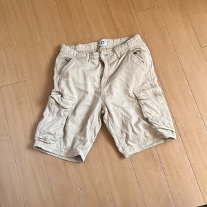 Beiga cargoshorts från 157, stl 160 - Säljer ett par beiga cargoshorts från 157 med flera stora fickor på sidorna och klassisk cargomodell. Shortsen har normal passform, bälteshällor och är gjorda i mjuk bomull. Perfekta för sommardagar när du vill ha en chill och praktisk stil.
