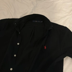 Svart skjorta från Ralph Lauren - Klassisk svart skjorta från Ralph Lauren med lång ärm, knappar framtill och röd broderad logga på bröstet. Oxford skjorta. Aldrig använd. Inga defekter. Pris kan diskuteras😁passar vid längd runt 170
