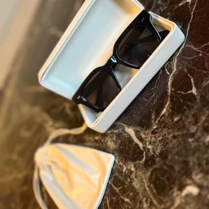 Svarta CHIMI solglasögon med etui - Snygga svarta solglasögon från CHIMI med fyrkantig form och breda bågar. Kommer med vitt hårt etui och mjuk påse. Glasen är mörka och bågarna har två små silverprickar på framsidan. Perfekt accessoar för att höja din stil.