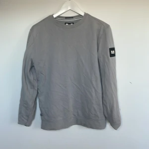 Grå sweatshirt från Weekend Offender XS - Säljer en stilren grå sweatshirt från Weekend Offender i 100% bomull. Tröjan har rund hals, lång ärm och en svart patch med logga på vänster ärm. Perfekt för dig som gillar enkel och clean streetstyle.