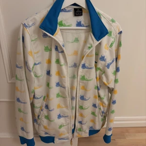 Jordan vit vindjacka med sneakersmönster - Vit vindjacka från Jordan med färgglatt sneakersmönster i blått, grönt och gult. Jackan har blå ribbade muddar och krage, hel dragkedja och två fickor fram. Perfekt för dig som gillar sportig och unik streetstyle.