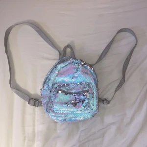 Glittrig miniryggsäck med paljetter - Superfin liten ryggsäck täckt av skimrande paljetter i blått, rosa och silver. Väskan har justerbara grå axelband, ett huvudfack och en mindre ficka framtill med dragkedja. Perfekt för dig som vill sticka ut med en färgglad och glittrig accessoar.
