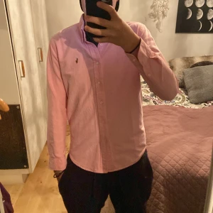 Rosa skjorta från Ralph Lauren M - Säljer en snygg rosa skjorta från Ralph Lauren i custom fit. Skjortan har klassisk krage, knappar hela vägen och broderad logga på bröstet. Tillverkad i bomull och har långa ärmar. Perfekt för dig som vill ha en stilren och fräsch look.