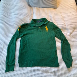 Grön långärmad pikétröja Polo Ralph Lauren - Grön långärmad pikétröja från Polo Ralph Lauren i slim fit. Stort gult broderat logotyp på bröstet och gul siffra 3 på en axeln. Passar dig som är runt 160, perfekt inför hösten/vintern.