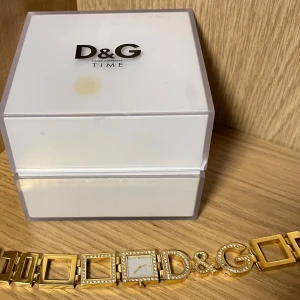 Guldig D&G armbandsklocka med kristaller - Snygg armbandsklocka från D&G i guldigt metall med fyrkantig urtavla och kristaller runt både urtavlan och länkarna. Klockan har dekorativa D&G-bokstäver och levereras i original ask. Perfekt statement piece för dig som gillar lyxig stil.
