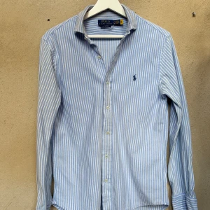 Blåvit randig skjorta Polo Ralph Lauren - Snygg blå och vit randig skjorta från Polo Ralph Lauren med klassisk krage, knappar framtill och broderad logga på bröstet. Tillverkad i mjuk bomull och perfekt för en clean och stilren look.