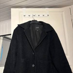 Svart kappa från Monki - Stilren svart kappa från Monki med klassisk krage och knäppning framtill. Jackan har en rak passform och är fodrad för extra värme. Perfekt för dig som vill ha en enkel men snygg look under kyliga dagar.
