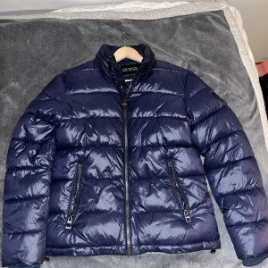 Blå pufferjacka från Guess - Snygg marinblå pufferjacka från Guess med hög krage och två dragkedjeförsedda fickor framtill. Jackan har quiltad design och är tillverkad i glansig polyester. Perfekt för kalla dagar och ger en clean streetstyle-look.