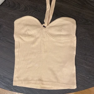 Beige ribbad halterneck korsett Gina Tricot - Supersnygg beige ribbad korsett-topp från Gina Tricot i halterneck-modell i storlek XXS. Toppen har markerade kupor och smala axelband som knyts i nacken. Perfekt för dig som gillar en tight och trendig look. Materialet är mjukt och stretchigt för bästa passform.
