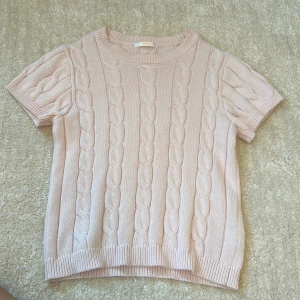 Ljusrosa kortstickad tröja från Gina Tricot - Supersöt ljusrosa stickad tröja från Gina Tricot med korta ärmar och klassiskt kabelstickat mönster. Tröjan har rund halsringning och ribbade kanter, perfekt för lager-på-lager eller sommarens svalare dagar.