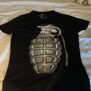 Svart t-shirt Horspist Paris XL - Svart t-shirt från Horspist Paris i storlek XL med en stor glittrig granat framtill. T-shirten har korta ärmar och är tillverkad i mjuk bomull. Snygg detalj med paljetter och metallmärke i nacken. Perfekt för dig som vill sticka ut.