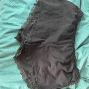 Svarta shorts med spets från H&M - Svarta shorts från H&M i mjuk bomull med elastisk midja och fina spetsdetaljer längs bensluten. Perfekta för en chill look eller som mysplagg hemma. Klassisk och enkel design med en feminin touch.