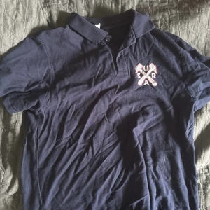 Mörkblå pikétröja Surahammar  - Snygg mörkblå pikétröja från SRA med korta ärmar och klassisk krage. På bröstet finns ett coolt tryck med korsade yxor och bokstäver. Tröjan är gjord i mjuk bomull och har knappar vid halsen för en schysst look.