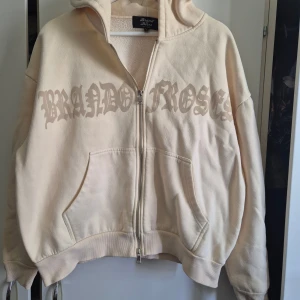 Gräddvit hoodie från Brand of Roses - Säljer en boxig, croppad hoodie från Brand of Roses gräddvit med stor logga över bröstet. Tröjan har dragkedja, huva och två stora fickor framtill. Tillverkad i mjuk bomullsfleece för extra comfort och värme. Perfekt för dig som gillar streetwear och snygga detaljer.