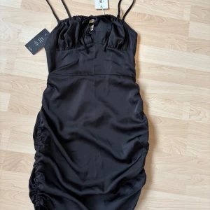 Svart satin klänning från NA-KD - Svart kortklänning i satin från NA-KD med smala axelband. Klänningen har en liten knytning framtill vid bysten och en figurnära passform. Oanvänd med lapp kvar på 