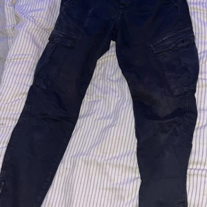 Mörkblå cargopants från Gabba - Säljer ett par mörkblå cargopants från Gabba med flera stora fickor på sidorna och bak. Byxorna har en tapered passform och är tillverkade i bomull. Perfekta för dig som gillar en avslappnad och funktionell stil.