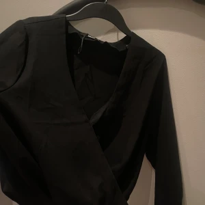 Svart omlottklänning från BikBok - Svart omlottklänning från BikBok i storlek L. Klänningen har lång ärm, v-ringning och knäppning i sidan. Stilren och enkel design i ett mjukt tyg som känns skönt mot huden. Perfekt för dig som gillar minimalistiska och klassiska plagg.