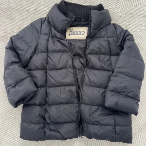Trekvartsärmad pufferjacka från Herno - Säljer en svart pufferjacka från Herno med hög krage och ribbad insida vid halsen. Jackan har quiltad design, dragkedja framtill och en loose passform med trekvartsärmar. Perfekt för kyliga dagar och enkel att styla till jeans eller hoodies.