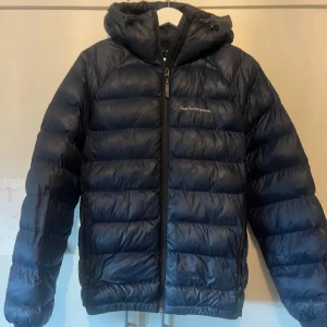 Mörkblå dunjacka Peak Performance S - Snygg mörkblå dunjacka från Peak Performance i storlek S. Jackan har huva, dragkedja framtill och klassisk pufferdesign med horisontella sömmar. Perfekt för kalla dagar och riktigt skön att bära. Logga på bröstet och praktiska fickor. OBS ytterst liten defekt på ungefär en halv cm som knappt syns vid armbågen och är jätte lätt att laga