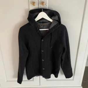 Svart hoodie-kofta från Massimo Dutti - Säljer en svart hoodie-kofta från Massimo Dutti i ullmix med silke. Tröjan har knäppning med knappar framtill, huva med snörning och långa ärmar. Snygg och enkel att matcha med det mesta. Den passar S-M
