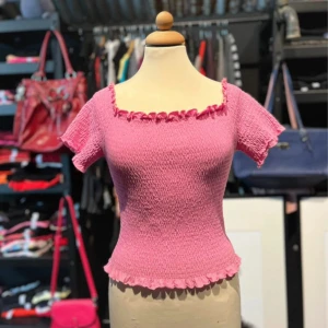 Rosa smockad topp med volangkant - Supersöt rosa topp med smockad stretch och volangdetaljer vid halsringning och nederkant. Toppen har korta ärmar och en fyrkantig urringning som ger en feminin vibe. Perfekt för dig som gillar färg och vill sticka ut lite extra.