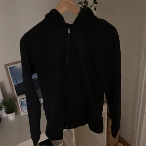 Mörkblå hoodie från Jack & Jones - Svart hoodie från Jack & Jones med dragkedja framtill och huva. Tröjan har långa ärmar, ribbade muddar och två fickor framtill. Perfekt för chill dagar eller när du vill ha en enkel och stilren look.