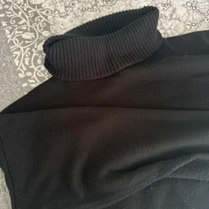 Svart polotröja i stickat material - Svart polotröja med ribbad hög krage och stickad känsla. Tröjan har klassisk passform och är perfekt för kyligare dagar. Enkel och stilren design som funkar till många olika outfits.