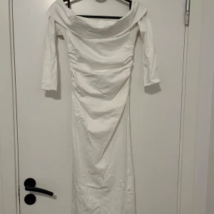 Vit långklänning från Boohoo - Säljer en elegant vit långklänning från Boohoo med offshoulder och trekvartsärm. Klänningen har snygga draperingar vid midjan och en bred krage som ger en cool och modern vibe. Perfekt för dig som vill sticka ut med stilren look.
