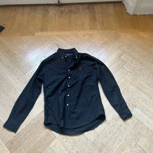 Svart skjorta från Ralph Lauren - Klassisk svart skjorta från Ralph Lauren med röd broderad logga på bröstet. Skjortan har lång ärm, knappar framtill och en snygg krage. Tillverkad i bomull och har en tidlös, normal passform. Perfekt för dig som gillar stilrena plagg med ikonisk touch.