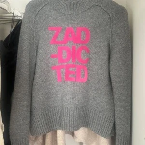 Grå stickad tröja med rosa text - Grå stickad tröja med hög hals och breda ribbade muddar. Framsidan har ett stort tryck i starkt rosa med texten 'ZADDICTED'. Perfekt för dig som gillar statement-plagg och vill sticka ut med färg och attityd. Barnstorlek men passar mig med xs-s