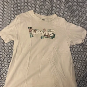 Vit t-shirt med tryck från Levi's - Vit t-shirt från Levi's med tropiskt inspirerat tryck över loggan på bröstet. Klassisk passform med rund hals och korta ärmar. Tillverkad i mjuk bomull, perfekt för en chill och stilren look.