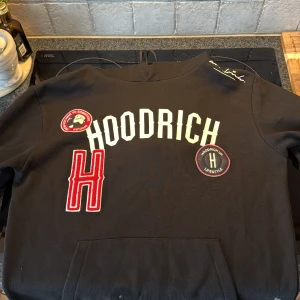 Svart hoodie från Hoodrich med patchar - Svart hoodie från Hoodrich i bomull med stora vita bokstäver och röda patchar framtill. Snygg broderad H och coola detaljer som dragsko i huvan. Perfekt streetstyle-plagg för dig som gillar att sticka ut.