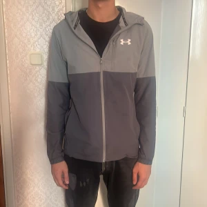  Under Armour windbreaker - Säljer en snygg tvåfärgad grå vindjacka från Under Armour med huva och dragkedja hela vägen. Jackan har en sportig look, elastiska muddar och logga på bröstet. Perfekt för dig som gillar stilren och funktionell design.