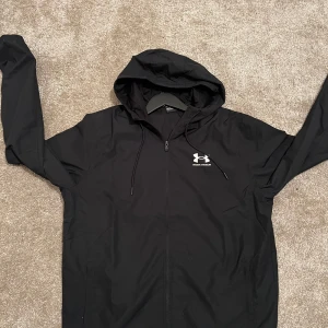 Svart vindjacka från Under Armour - Svart vindjacka från Under Armour med huva och dragsko. Perfekt för detta väder. Den är storlek M men kan även passa mig. Den passar mig bra som 179 cm 65 kg. Pris kan diskuteras litegrann. Använts ett fåtal gånger