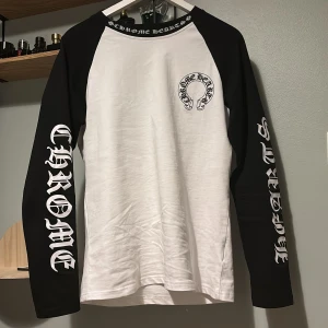 Chrome Hearts svartvit långärmad tröja - Säljer en långärmad svartvit tröja från Chrome Hearts med kontrastärmar och stora tryck på både ärmar och rygg. Tröjan har klassisk logga på bröstet och cool text längs ärmarna. Perfekt för dig som gillar streetwear och vill sticka ut.