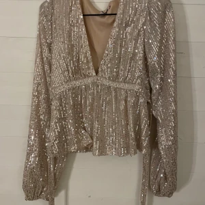 Glittrig paljettopp från Nelly - Supersnygg glittrig topp från Nelly i beige med massor av paljetter. Djup v-ringning framtill, långa puffiga ärmar och markerad midja. Perfekt för dig som vill sticka ut med en festlig look. Toppen har en croppad passform och är täckt av skimrande paljetter.