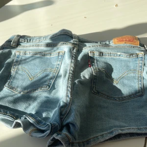 Levi's ljusblå jeansshorts - Säljer ett par klassiska ljusblå jeansshorts från Levi's med fem fickor, synliga knappar framtill och uppvikta benslut. Shortsen har den ikoniska läderpatchen bak och snygga kontrastsömmar. Perfekta för varma dagar och en avslappnad stil.