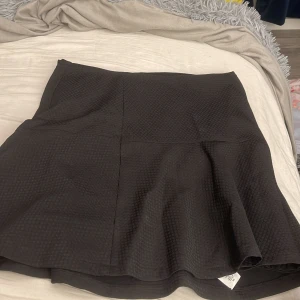 Svart kjol från Zoul Edition - Säljer en svart kortkjol från Zoul Edition i storlek S. Kjolen har en lätt utsvängd form och ett diskret rutmönster i tyget som ger en cool struktur. Perfekt att styla med en stickad tröja eller en snygg topp.