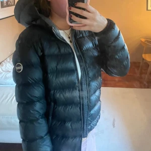Svart Colmar dunjacka med huva - Säljer en svart dunjacka från Colmar med huva och dragkedja. Jackan är quiltad och har en klassisk pufferstil med diskret logga på ärmen. Perfekt för kalla dagar!