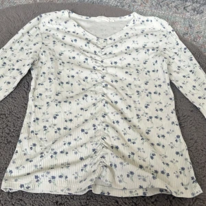 Blommig vit långärmad blus från KappAhl storlek 158/164 - Supersöt vit blus från KappAhl med blå blommor och diskreta ränder. Den har v-ringning, lång ärm och snygg snörning framtill som ger en cool rynkad effekt. Materialet är mjukt och känns som bomull, perfekt för vår och sommar. Använde en gång men tvättat fler 