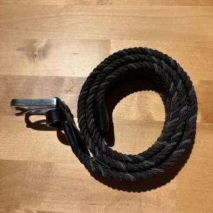 Flätat bälte från ONEWear Sweden - Snyggt flätat bälte från ONEWear Sweden i mörka nyanser av svart, grått och brunt. Bältet har detaljer i svart skinn och ett silvrigt spänne. Perfekt accessoar för att lyfta din outfit med en cool och stilren touch.