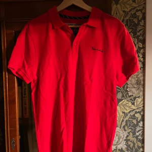 Röd pikétröja från Bondelid - Snygg röd pikétröja från Bondelid med klassisk krage och knappar framtill. Tröjan har korta ärmar och broderad logga på bröstet. Perfekt för en stilren och avslappnad look.