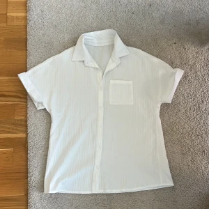 Vit kortärmad skjorta med ficka på med bra kvalitè och kondition - Vit skjorta med korta ärmar och klassisk krage. Skjortan har en bröstficka och knäppning framtill. Materialet är lätt och har en diskret struktur med tunna vertikala ränder. Perfekt för en clean och fräsch look.