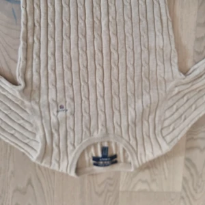 Beige kabelstickad tröja från GANT - Kabelstickad beige tröja från GANT i bomull. Klassisk rund halsringning, långärmad och med broderad logga på bröstet. Perfekt för lager-på-lager och snygg till både jeans och kjol.