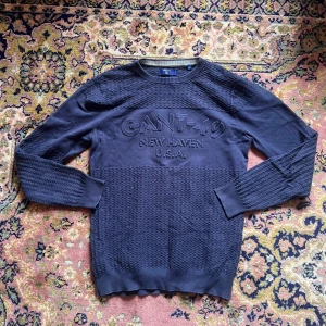Mörkblå stickad tröja från GANT M - Säljer en mörkblå stickad tröja från GANT med broderad logga och texten 'GANT-49 NEW HAVEN U.S.A.' framtill. Tröjan har rund hals, långärmade ärmar och ett snyggt strukturerat mönster på både ärmar och överkropp. Perfekt för dig som gillar klassisk och stilren design.