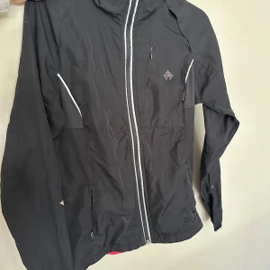 Svart vindjacka med vita detaljer - Svart vindjacka från Peak Performance med vita dragkedjor och detaljer. Jackan har hög krage, långa ärmar och praktiska fickor med dragkedja. Materialet är lätt och passar perfekt för blåsiga dagar. Snygg logga på bröstet och paneler i stretchigt tyg för extra komfort.